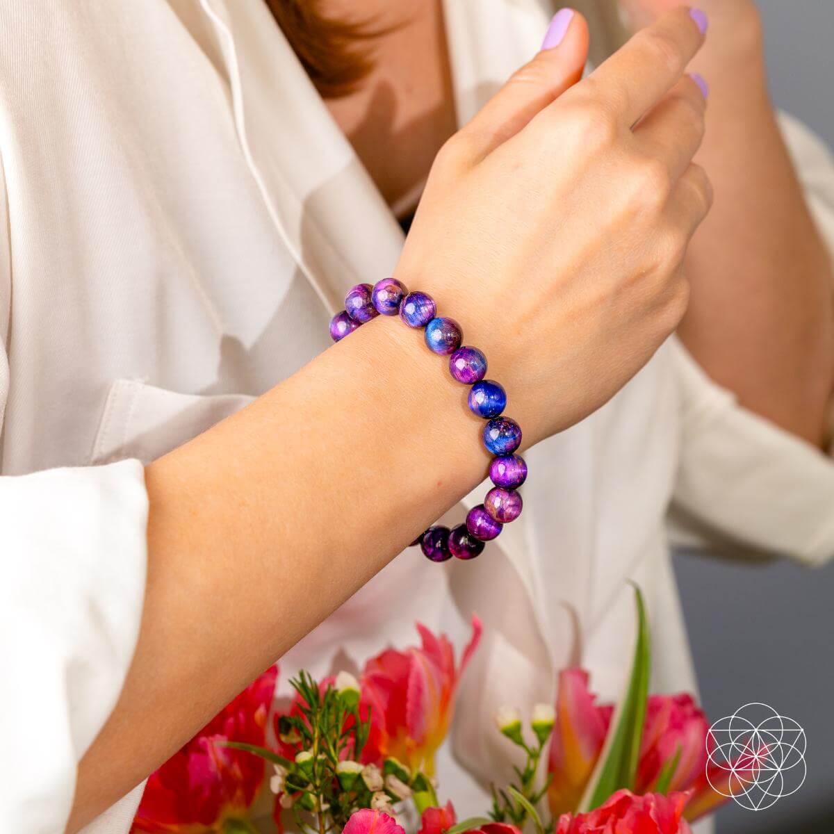 A person in a white shirt shows off the Destiny Activator - All-Galaxy Tiger’s Eye Bracelet by Conscious Items, featuring purple and blue marbled beads, while holding pink and red flowers, suggesting a touch of cosmic destiny.