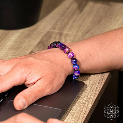 Someone types on a laptop at a wooden desk, wearing the Destiny Activator - All-Galaxy Tiger’s Eye Bracelet by Conscious Items.
