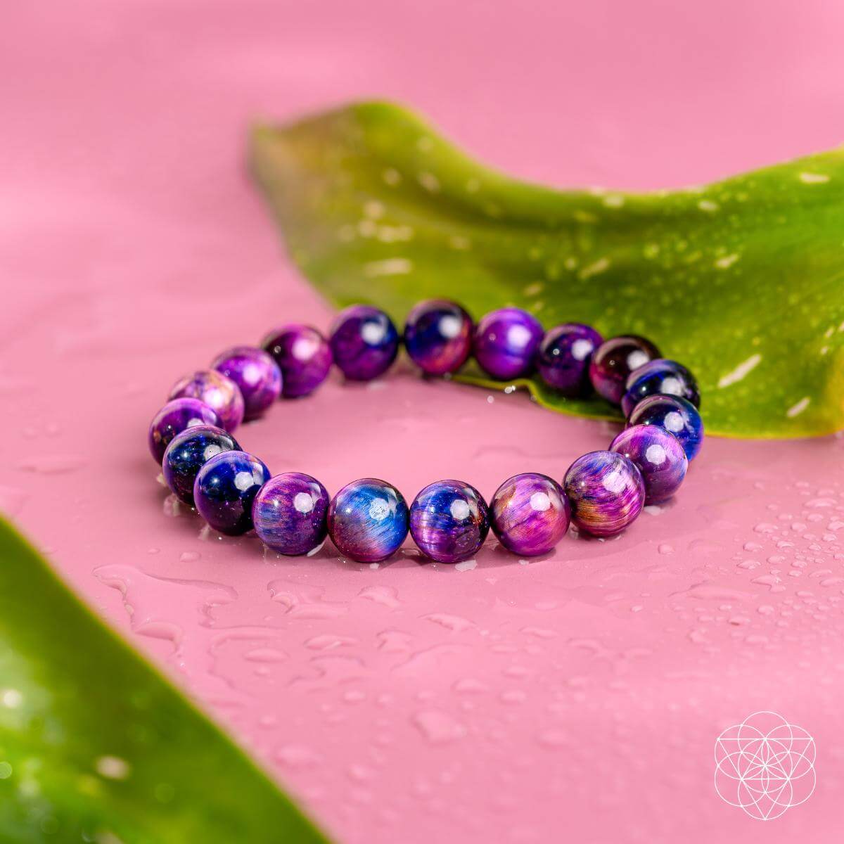 The Destiny Activator - All-Galaxy Tiger’s Eye Bracelet by Conscious Items, featuring shiny purple, blue, and pink beads, rests on a wet pink surface with two green leaves in the background.