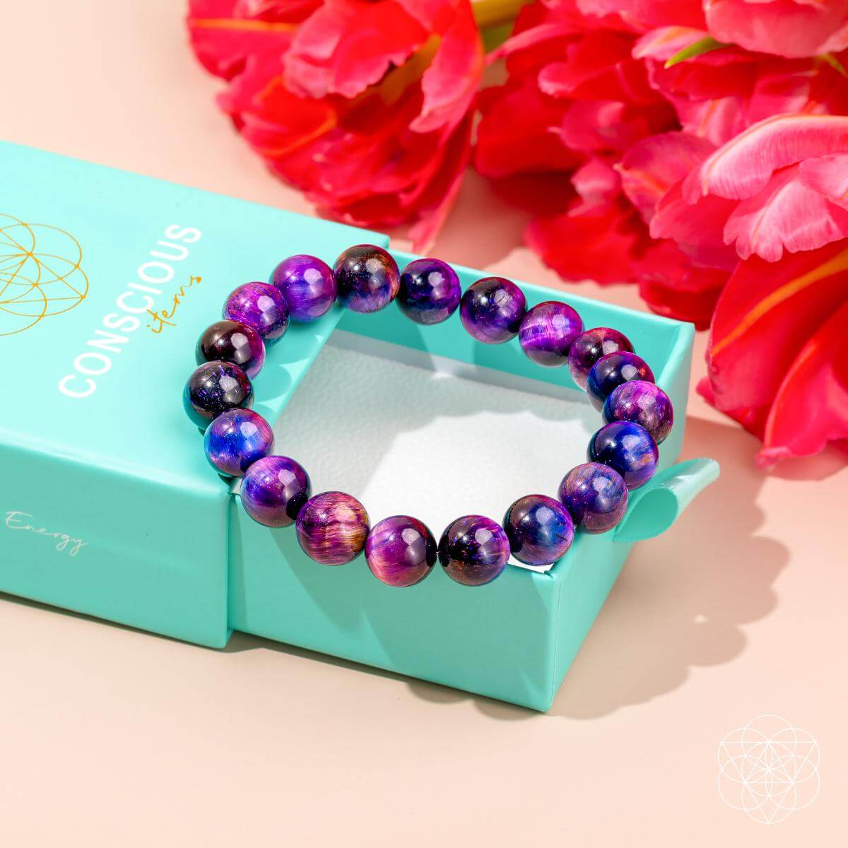 The Destiny Activator - All-Galaxy Tiger’s Eye Bracelet by Conscious Items, featuring purple and blue stones, is shown on an open turquoise Conscious Items box with bright red and pink flowers in the background.