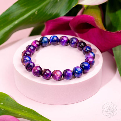 The Destiny Activator - All-Galaxy Tiger’s Eye Bracelet from Conscious Items features glossy purple, blue, and pink stones, displayed on a round white stand with green leaves and pink flowers against a light pink background.