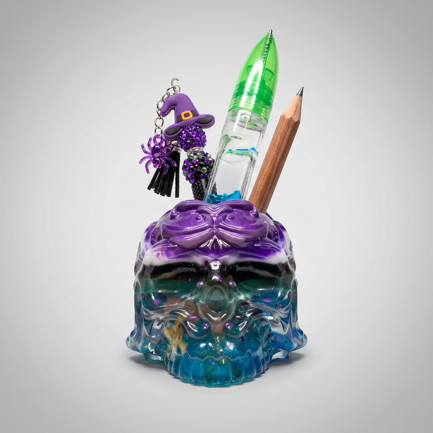 The Dragonsteins Monster Resin Skull by GorgeousDragon Designs LLC. is a colorful, iridescent desk organizer holding a pencil, green and clear pen, blue USB drive, and a keychain with a purple witch hat and beaded spider on a light gray background.