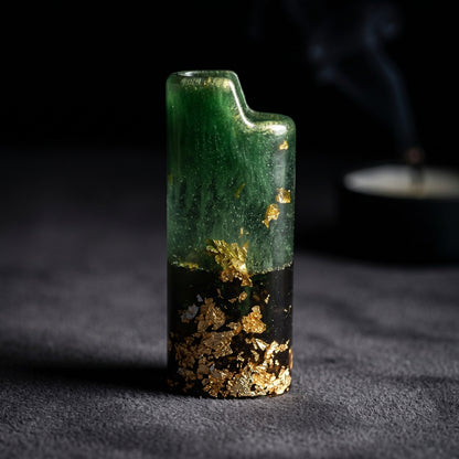 Handcrafted resin lighter case with emerald green swirls, a black base, and embedded gold leaf flakes on a metal pedestal.
