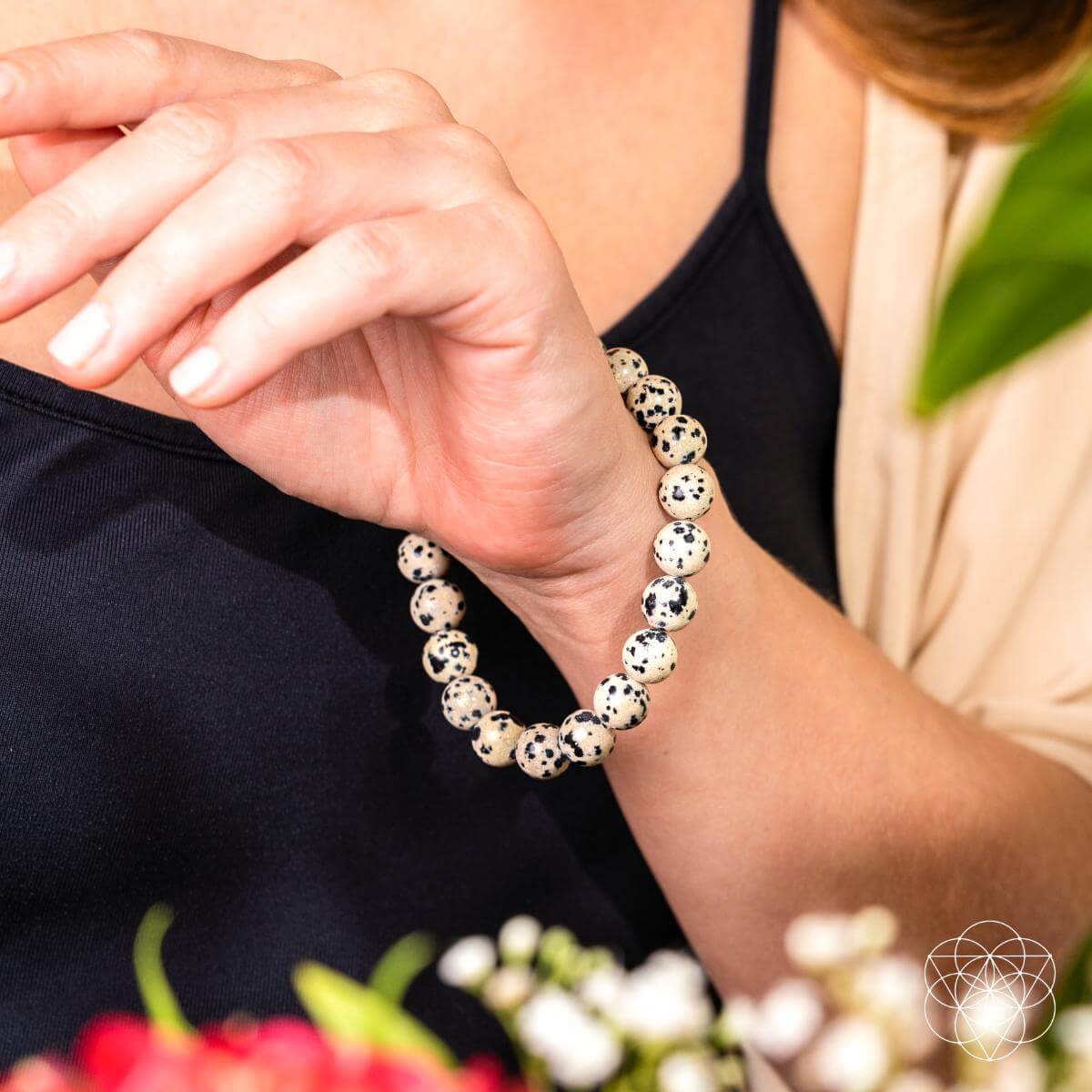 A person wearing the Emotional Detox - Dalmatian Jasper Bracelet by Conscious Items rests their hand near their face. Dressed in a black top and beige cardigan, they’re surrounded by blurred flowers and greenery, evoking emotional balance.