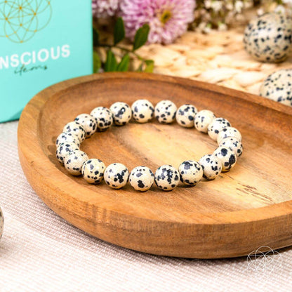 The Emotional Detox - Dalmatian Jasper Bracelet by Conscious Items is shown on a wooden dish, with its turquoise box and flowers in the background. The cream beads feature unique black spots and are known for supporting emotional balance.