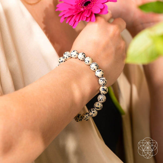 A person in a cream top holds a pink daisy, wearing the Emotional Detox - Dalmatian Jasper Bracelet by Conscious Items, believed to support emotional balance. Sunlight highlights their arm as green leaves appear in the foreground.