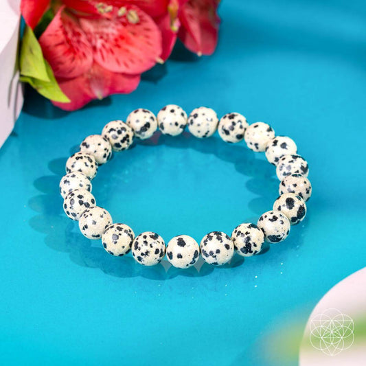 The Emotional Detox - Dalmatian Jasper Bracelet by Conscious Items, featuring round white stones with black spots, is shown on a turquoise surface with pink flowers. This bracelet promotes emotional balance and energy cleansing.