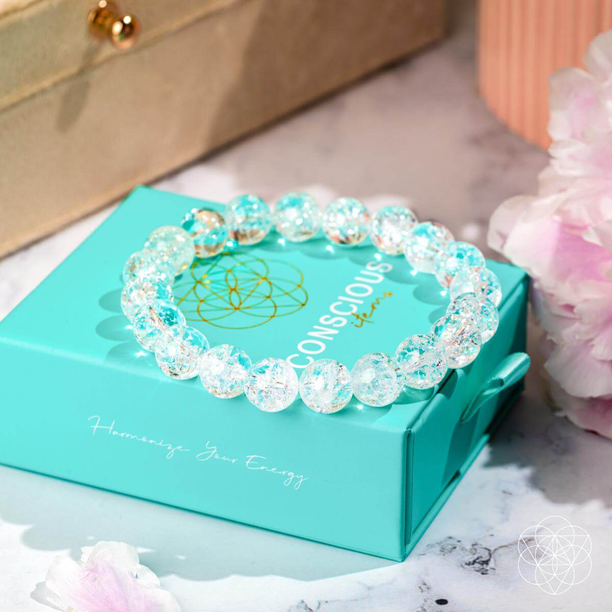 The Emotional Ice Bath – The Ice Quartz Bracelet by Conscious Items rests on a turquoise box, surrounded by soft pink flowers and a beige jewelry case, creating an elegant display that evokes emotional clarity.