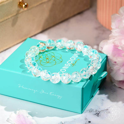 The Emotional Ice Bath – The Ice Quartz Bracelet by Conscious Items rests on a turquoise box, surrounded by soft pink flowers and a beige jewelry case, creating an elegant display that evokes emotional clarity.