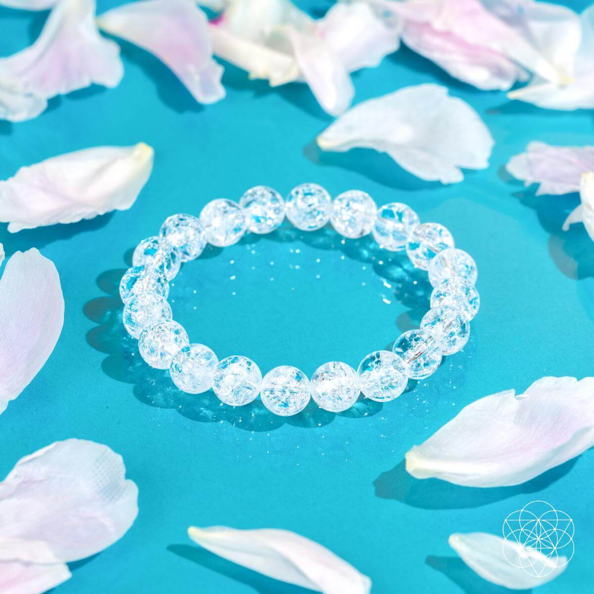 The Emotional Ice Bath – The Ice Quartz Bracelet from Conscious Items, a reset stone for emotional clarity, rests on a bright blue surface with pale pink and white petals. A white geometric logo is displayed in the bottom right corner.