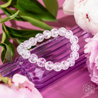 The Emotional Ice Bath – The Ice Quartz Bracelet by Conscious Items rests on a ribbed purple vase, surrounded by pink peonies and greenery, evoking emotional clarity. A geometric white logo appears in the bottom right corner.