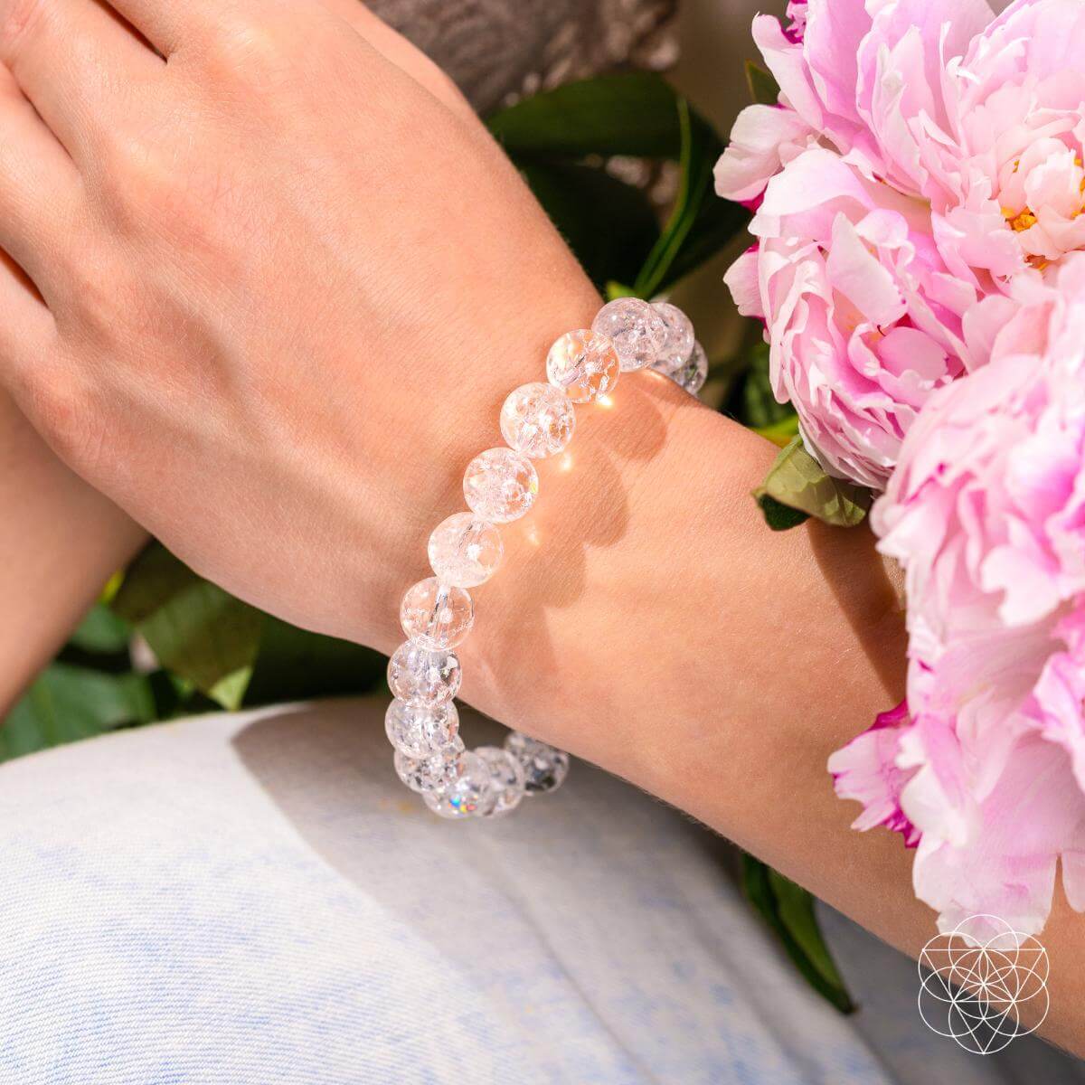 A hand adorned with the Emotional Ice Bath – The Ice Quartz Bracelet by Conscious Items rests near pink peonies, with natural light enhancing the flowers and jewelry, reflecting the bracelet's promise of emotional clarity and tranquility.
