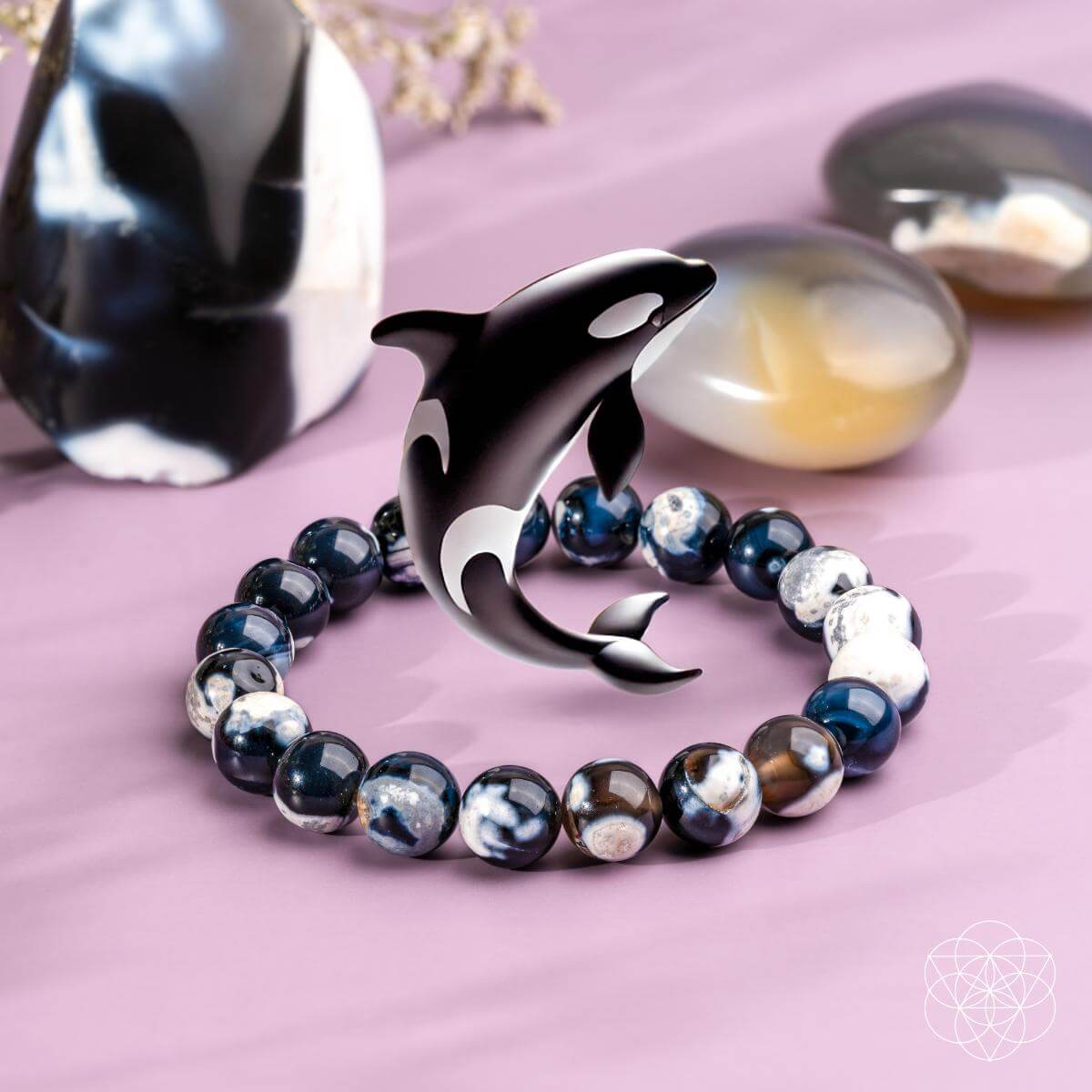 The Exotic Orca Agate Bracelet of Deep Healing by Conscious Items—adorned with Madagascar crystal and a metallic orca dolphin charm—rests on a pink surface amid polished stones, radiating soothing ocean energy gemstone vibes.