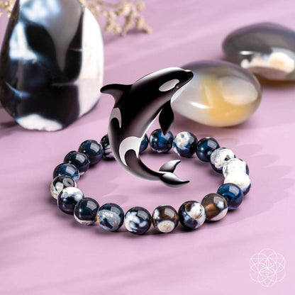 The Exotic Orca Agate Bracelet of Deep Healing by Conscious Items—adorned with Madagascar crystal and a metallic orca dolphin charm—rests on a pink surface amid polished stones, radiating soothing ocean energy gemstone vibes.