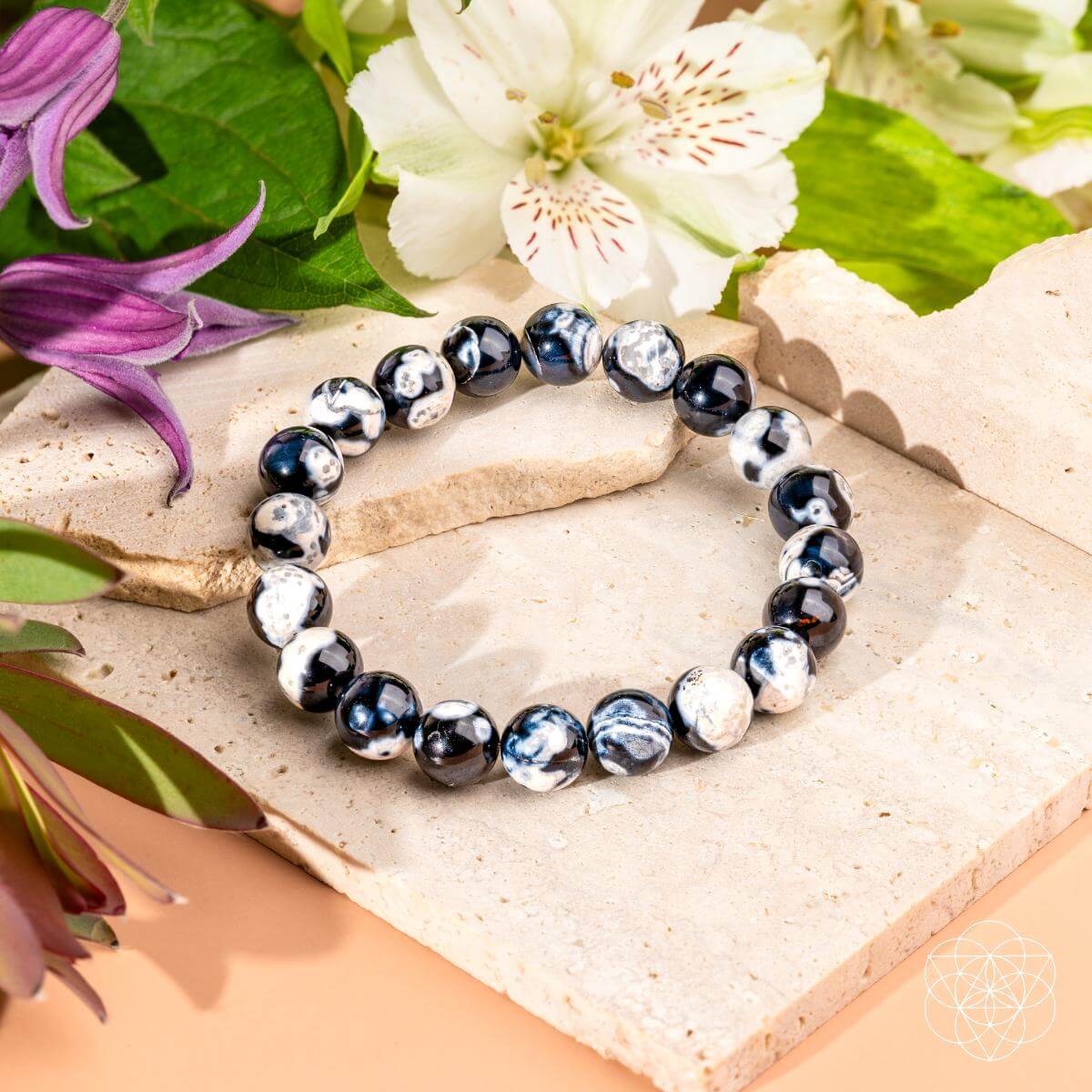 The Exotic Orca Agate Bracelet of Deep Healing by Conscious Items, featuring black, white, and blue marbled stones, is displayed on beige tiles among green leaves and white and purple flowers.