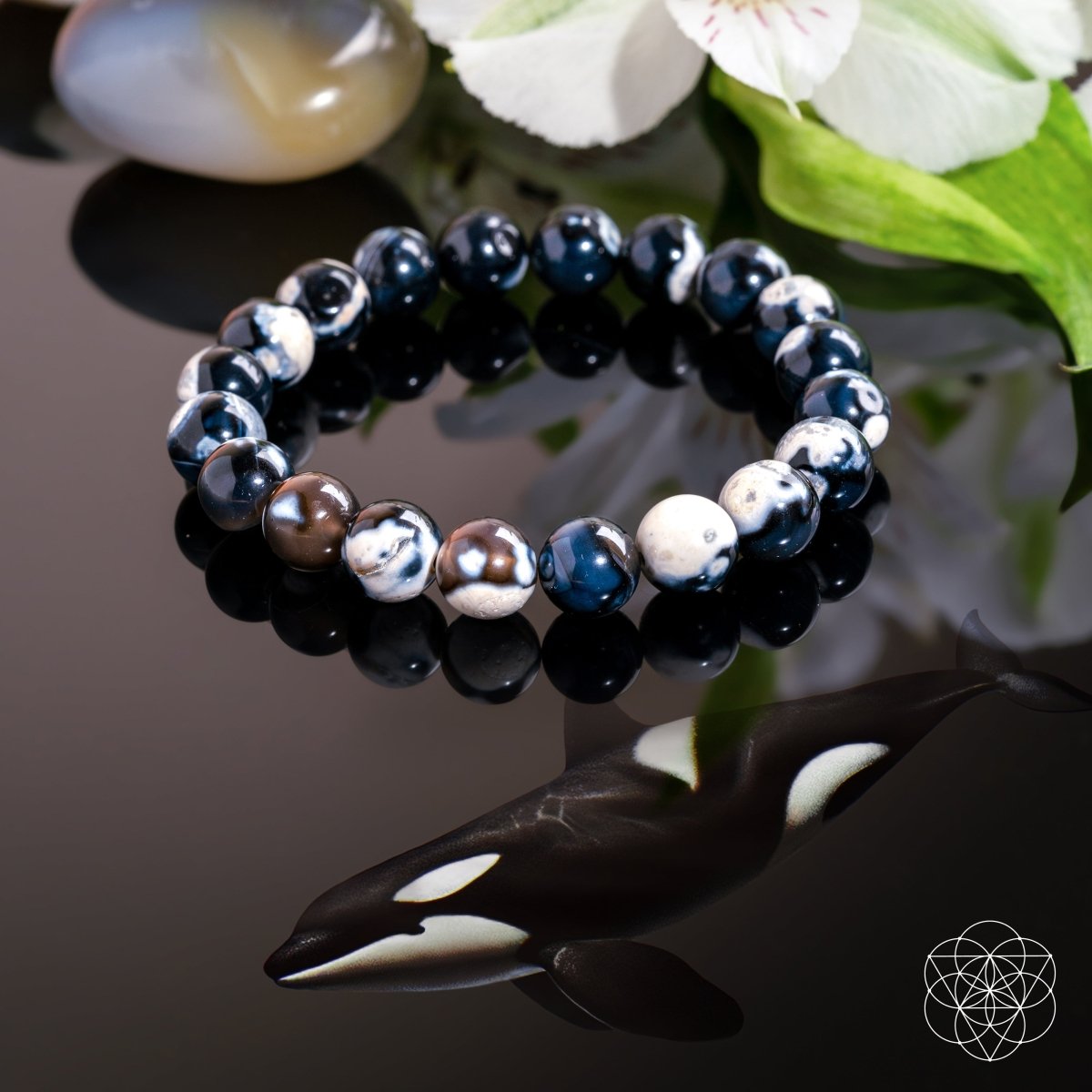 The Exotic Orca Agate Bracelet of Deep Healing by Conscious Items features black, white, and marbled stones on a glossy black surface reflected like an orca, with green leaves, a white flower, a Madagascar crystal, and a geometric logo nearby.