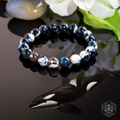 The Exotic Orca Agate Bracelet of Deep Healing by Conscious Items features black, white, and marbled stones on a glossy black surface reflected like an orca, with green leaves, a white flower, a Madagascar crystal, and a geometric logo nearby.