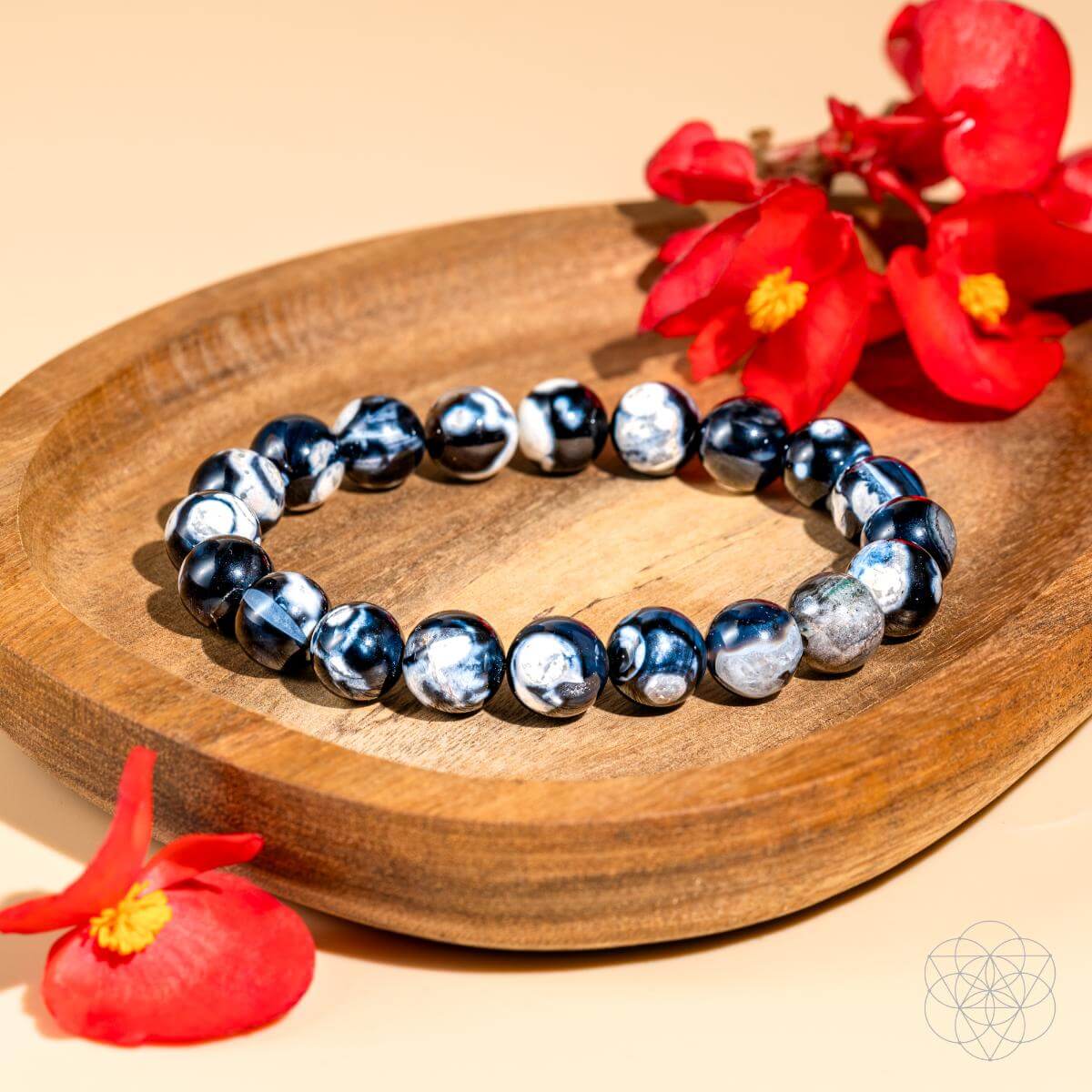 The Exotic Orca Agate Bracelet of Deep Healing by Conscious Items sits on a wooden dish with vivid red flowers nearby, on a beige surface featuring a subtle geometric design in the bottom right corner.