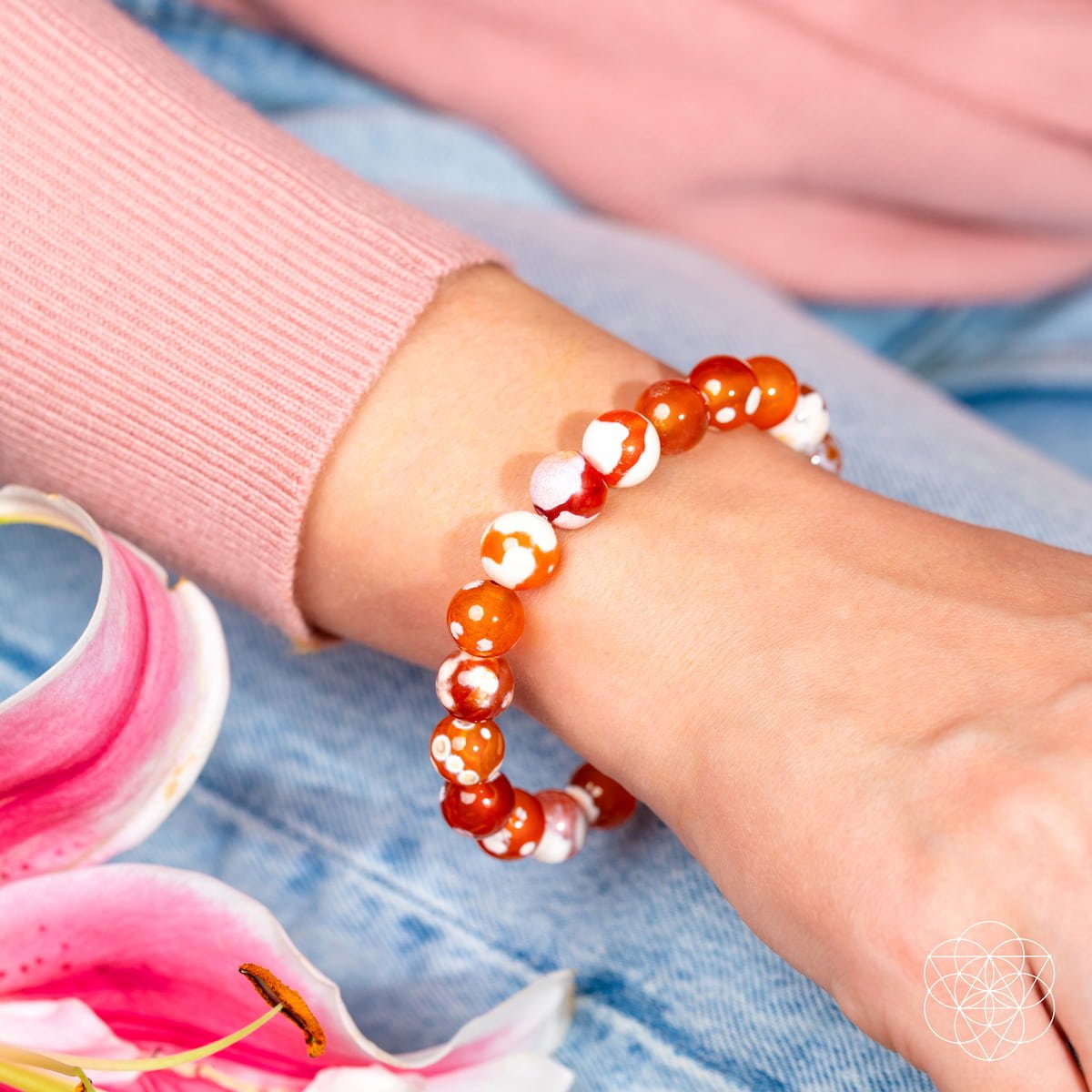 A close-up shows a wrist adorned with the Conscious Items Feminine Force – Carnelian Orca Agate Bracelet, featuring orange and white stones. The person wears a pink sweater and blue jeans, with pink flowers in the foreground—a chic bracelet for women.