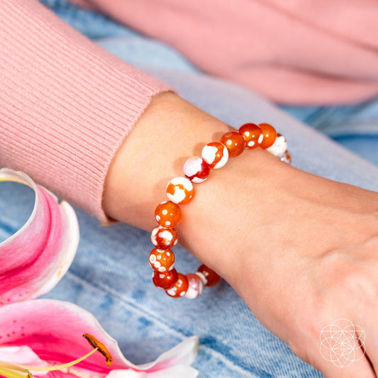 A close-up shows a wrist adorned with the Conscious Items Feminine Force – Carnelian Orca Agate Bracelet, featuring orange and white stones. The person wears a pink sweater and blue jeans, with pink flowers in the foreground—a chic bracelet for women.