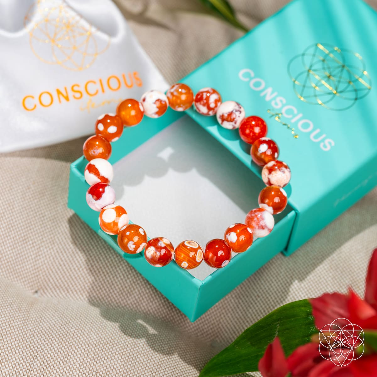 The Feminine Force – Carnelian Orca Agate Bracelet by Conscious Items rests on a turquoise branded box with its pouch and red flowers, set on light fabric—an elegant women’s bracelet.