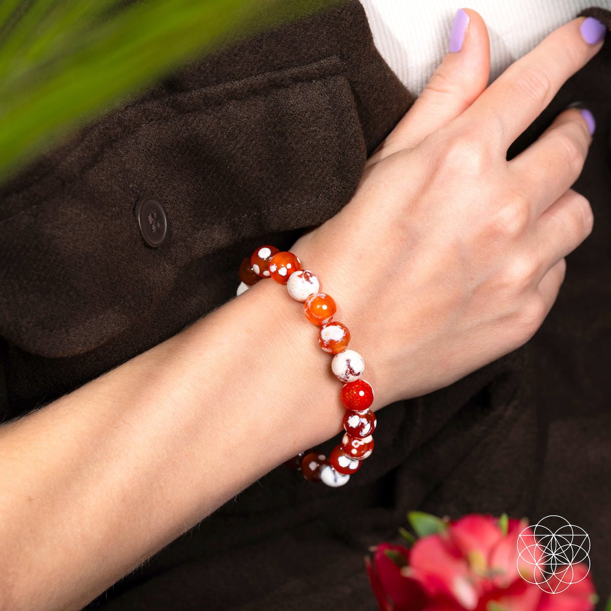 A hand with light purple nail polish rests on brown fabric, wearing the Feminine Force – Carnelian Orca Agate Bracelet by Conscious Items. Pink flowers are visible in the lower corner.