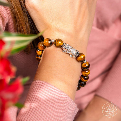 Wearing the Fortune Flows - Tiger’s Eye & Pixiu Bracelet by Conscious Items, paired with a pink top and surrounded by blurred red flowers, inspires Feng Shui abundance.