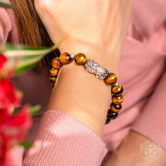 Wearing the Fortune Flows - Tiger’s Eye & Pixiu Bracelet by Conscious Items, paired with a pink top and surrounded by blurred red flowers, inspires Feng Shui abundance.