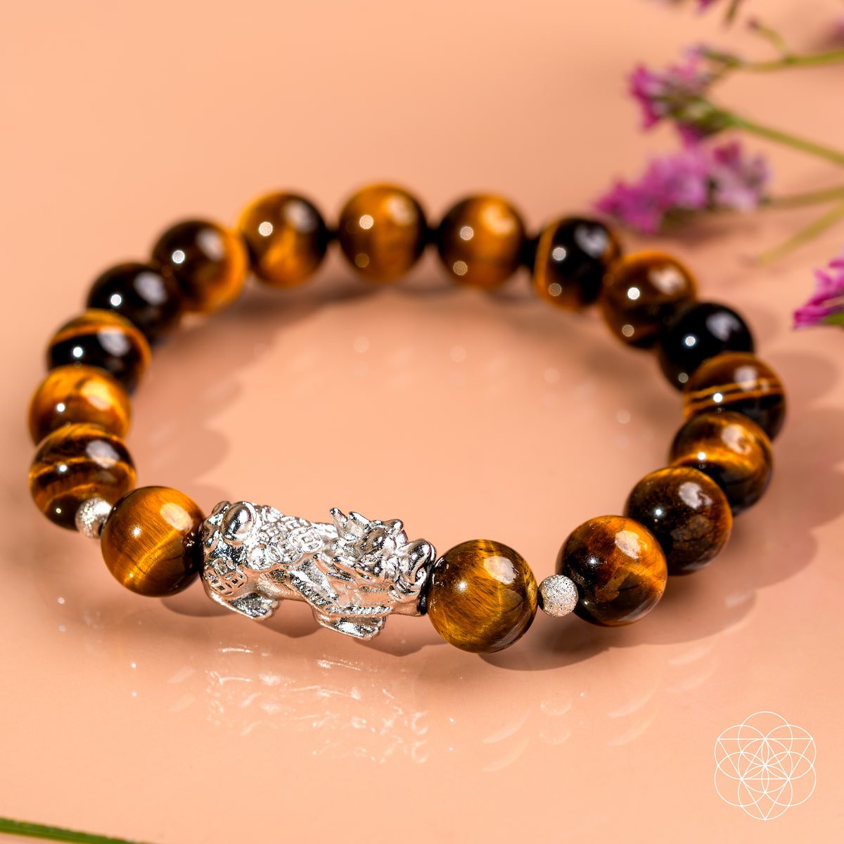The Fortune Flows - Tiger’s Eye & Pixiu Bracelet by Conscious Items, shown with polished beads and a silver Pixiu charm, is displayed on a peach surface near purple flowers and is said to attract abundance and positive energy.
