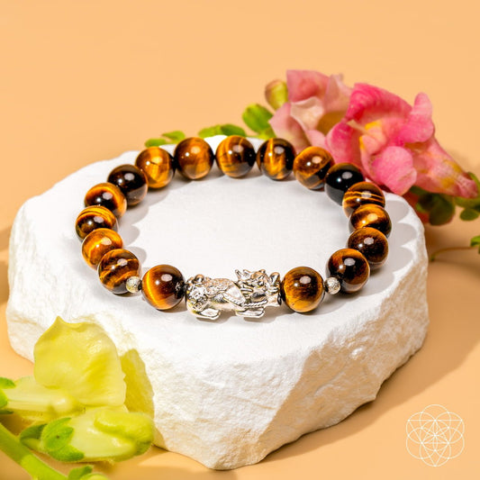 The Fortune Flows - Tiger’s Eye & Pixiu Bracelet by Conscious Items, showcased on a white stone with pink and yellow blooms, channels Feng Shui abundance against a calm pale yellow backdrop.