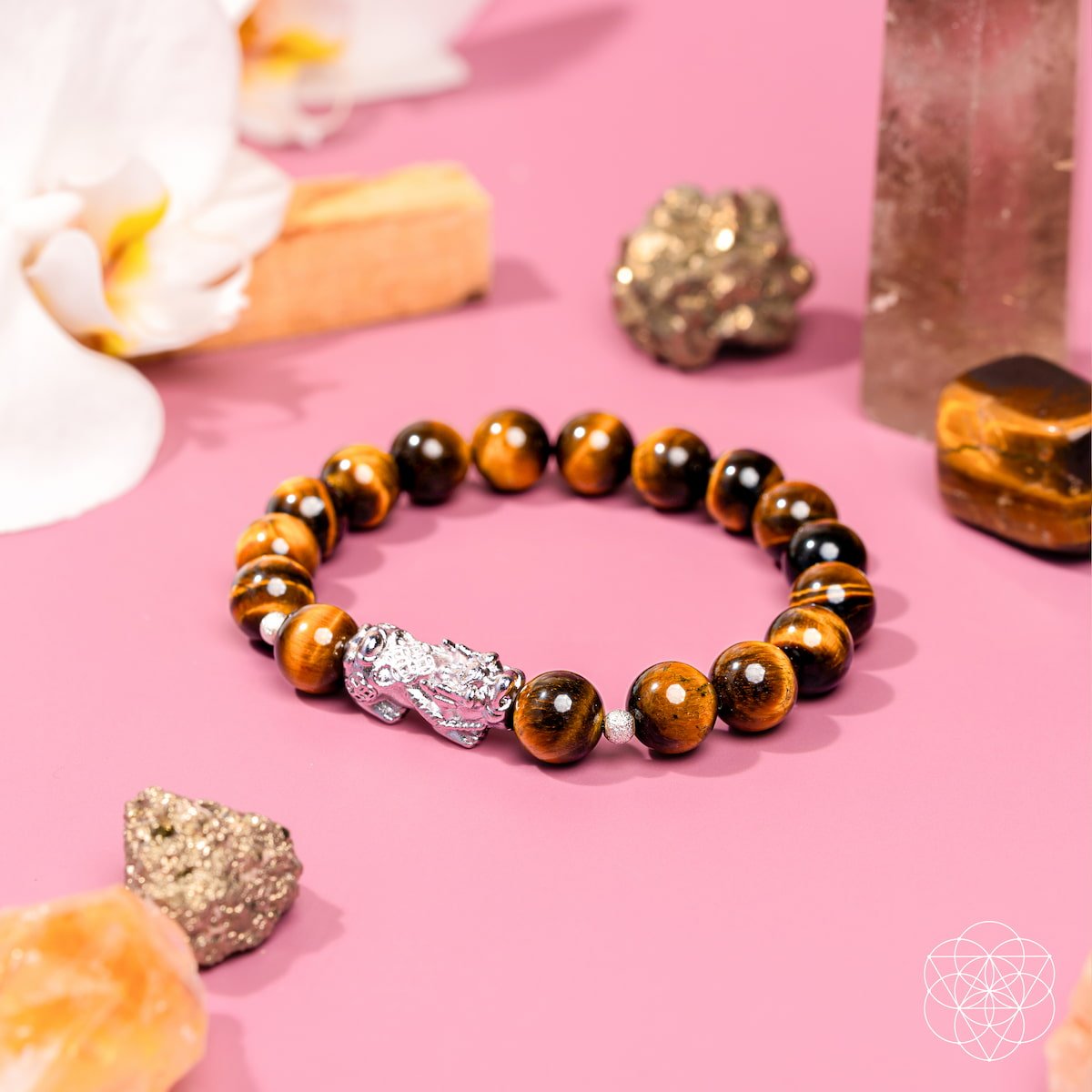 The Fortune Flows - Tiger’s Eye & Pixiu Bracelet by Conscious Items rests on a pink surface, encircled by crystals, minerals, and white flowers—an elegant Feng Shui-inspired piece for abundance and prosperity.