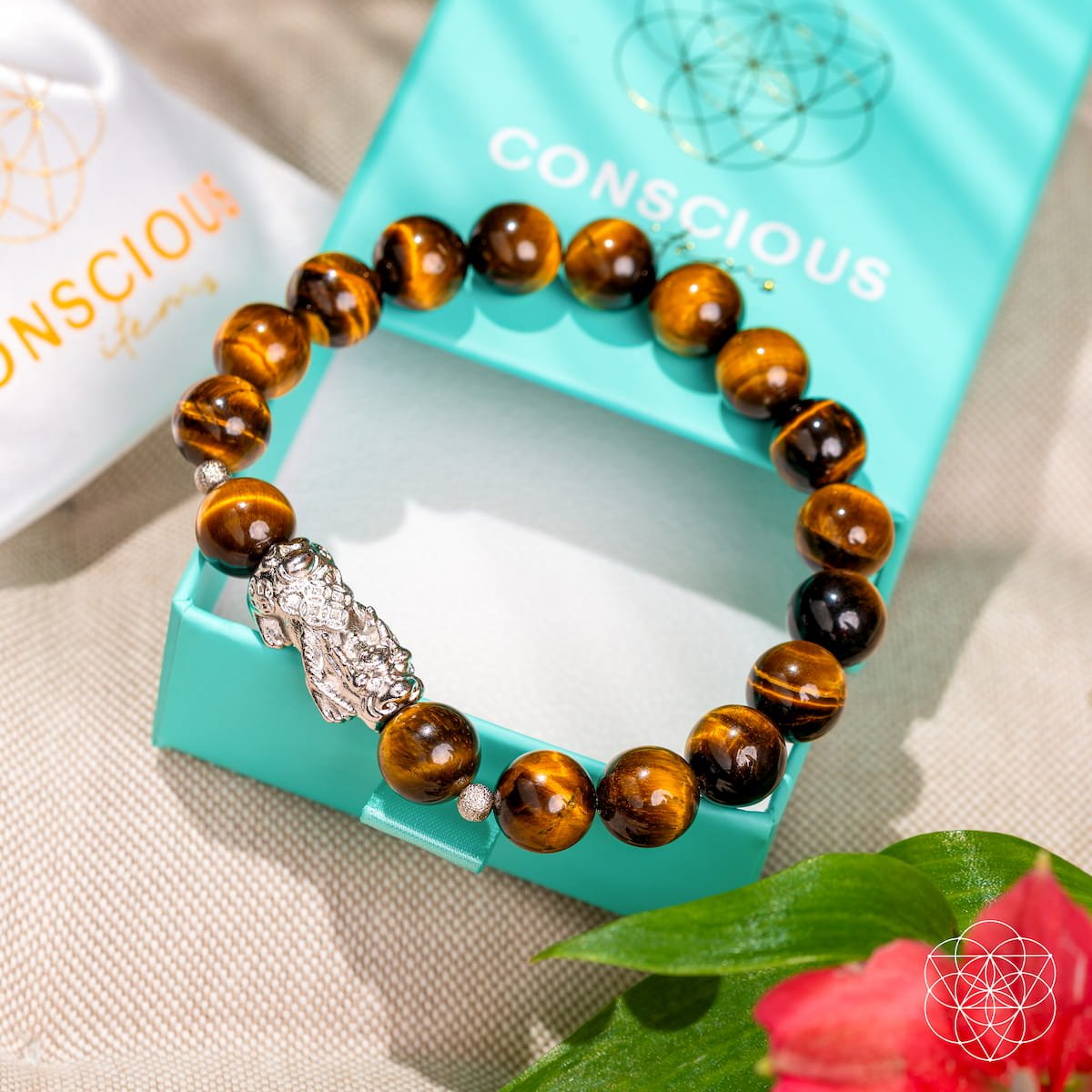 The Fortune Flows - Tiger’s Eye & Pixiu Bracelet by Conscious Items sits on an open turquoise branded box beside a pouch, with a red flower in the foreground.