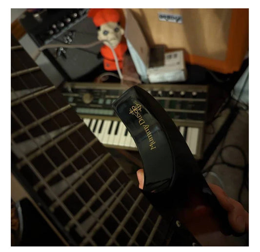 A hand holds a black Gorgeous Drag0n Mummy Dust guitar capo, with a guitar neck, keyboard, audio equipment, and red doll in the background.
