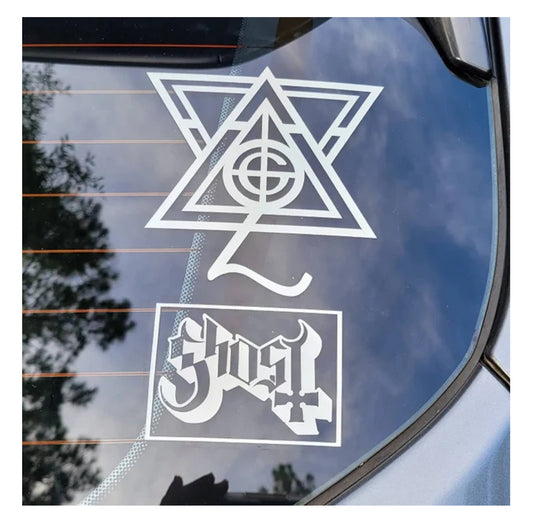 A car window displays two premium vinyl Papa Ghouls decals by Gorgeous Drag0n: one with a geometric triangle and circle, the other with “Ghost” in a gothic font, set against reflected trees and sky.