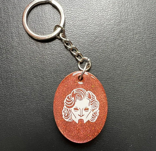 Keychain with a red oval pendant featuring a white gouleh mask design on a dark gray background