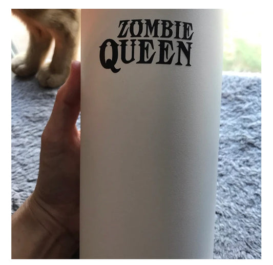 A hand holds the Zombie Queen white tumbler with bold black lettering, while a cat’s paws rest on a gray surface by the window in the background.