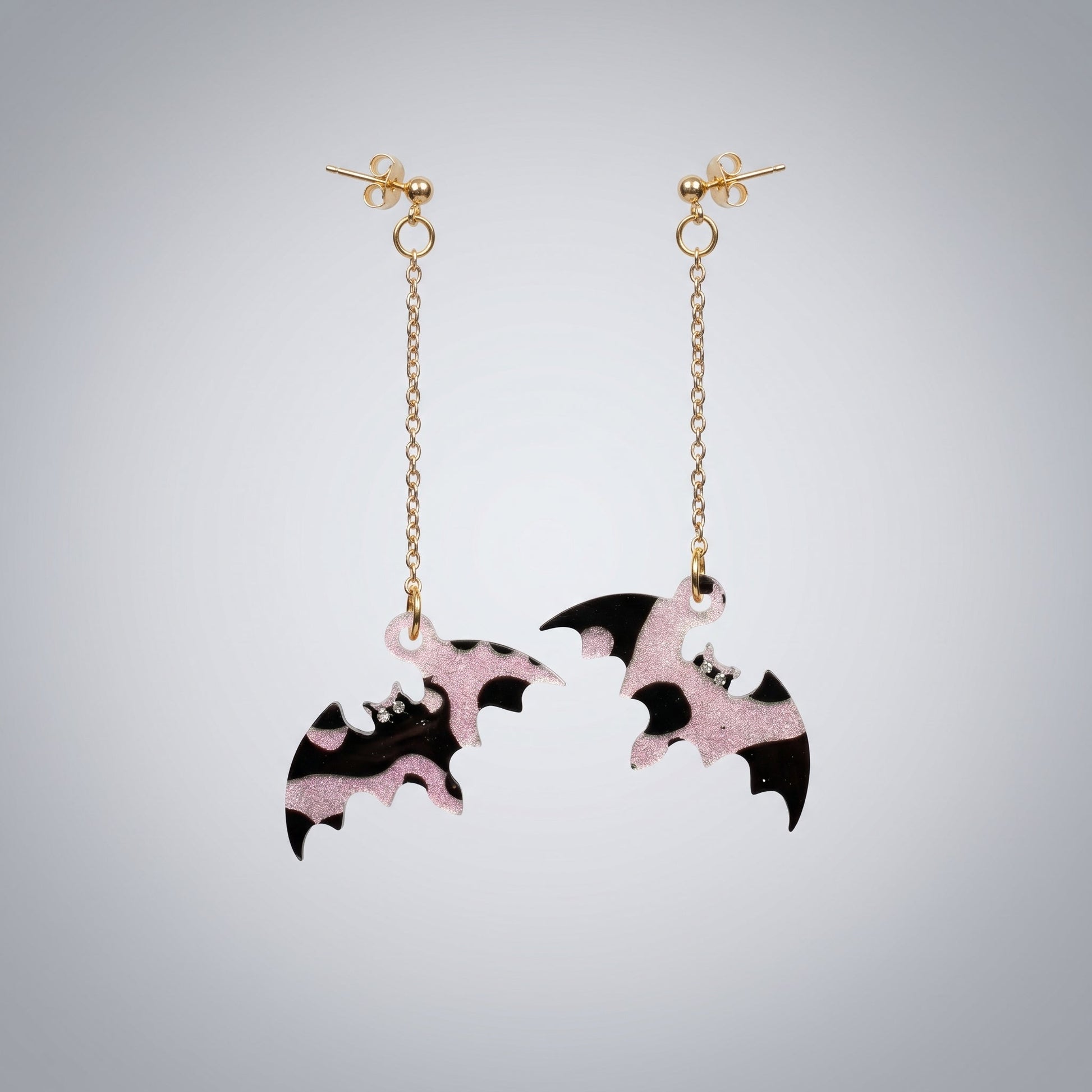 Dangle earrings featuring black and pink glitter resin bats with gold chains on a dark brushed metal surface.