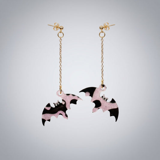 Dangle earrings featuring black and pink glitter resin bats with gold chains on a dark brushed metal surface.