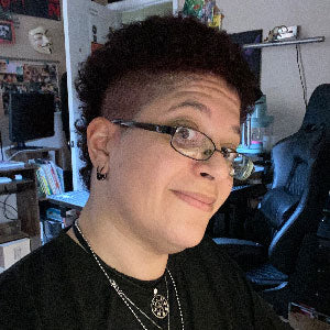 A person with short curly hair and glasses smiles at the camera. They are wearing earrings, necklaces, and a black shirt. The background shows a room with books, posters, and a large chair.