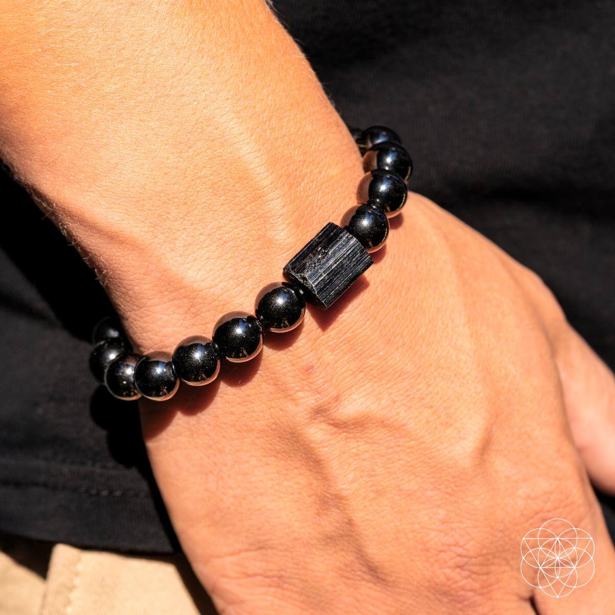 A close-up shows a wrist adorned with the Habit Breaker - Black Obsidian & Tourmaline Bracelet by Conscious Items, featuring a rectangular Black Obsidian centerpiece. The person wears a black shirt and beige pants.