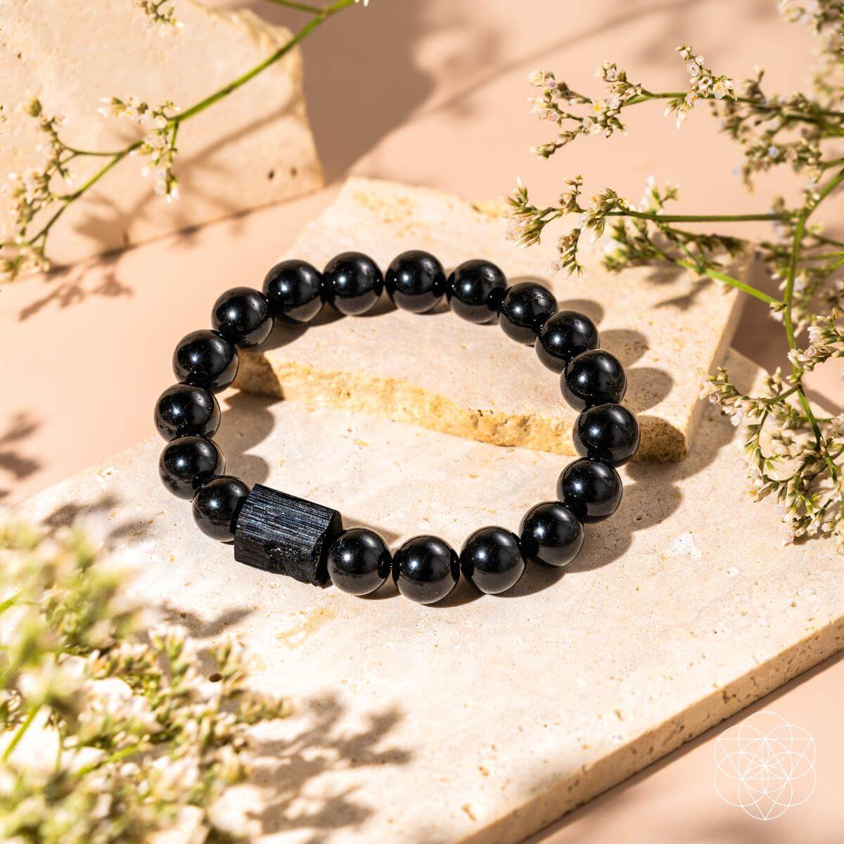 The Habit Breaker - Black Obsidian & Tourmaline Bracelet by Conscious Items is showcased on beige stone tiles, featuring a bold black obsidian bead and accented by delicate white flowers atop a light background.