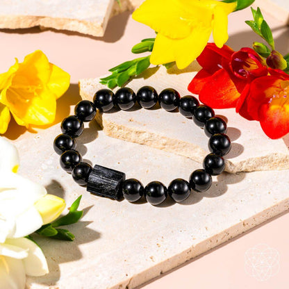 The Habit Breaker - Black Obsidian & Tourmaline Bracelet by Conscious Items is displayed on a light stone surface, surrounded by yellow, red, and white flowers. The bracelet features striking black beads with a large rectangular Black Obsidian accent.