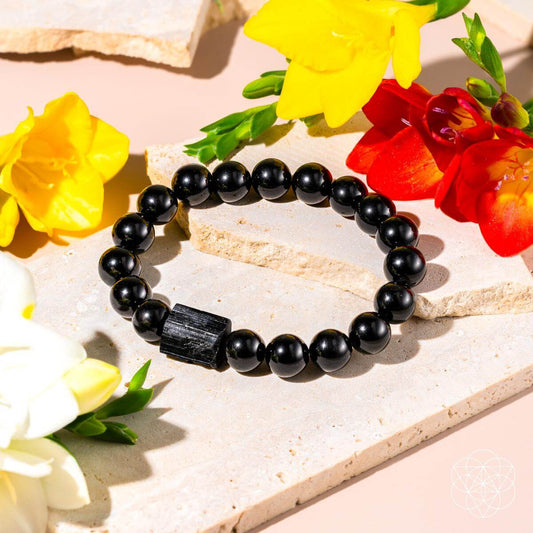 The Habit Breaker - Black Obsidian & Tourmaline Bracelet by Conscious Items is displayed on a light stone surface, surrounded by yellow, red, and white flowers. The bracelet features striking black beads with a large rectangular Black Obsidian accent.
