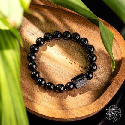The Habit Breaker - Black Obsidian & Tourmaline Bracelet from Conscious Items rests on a wooden dish with green leaves and natural light. Its rectangular charm and sleek black beads embody style and purpose, with a geometric white logo in the bottom right corner.