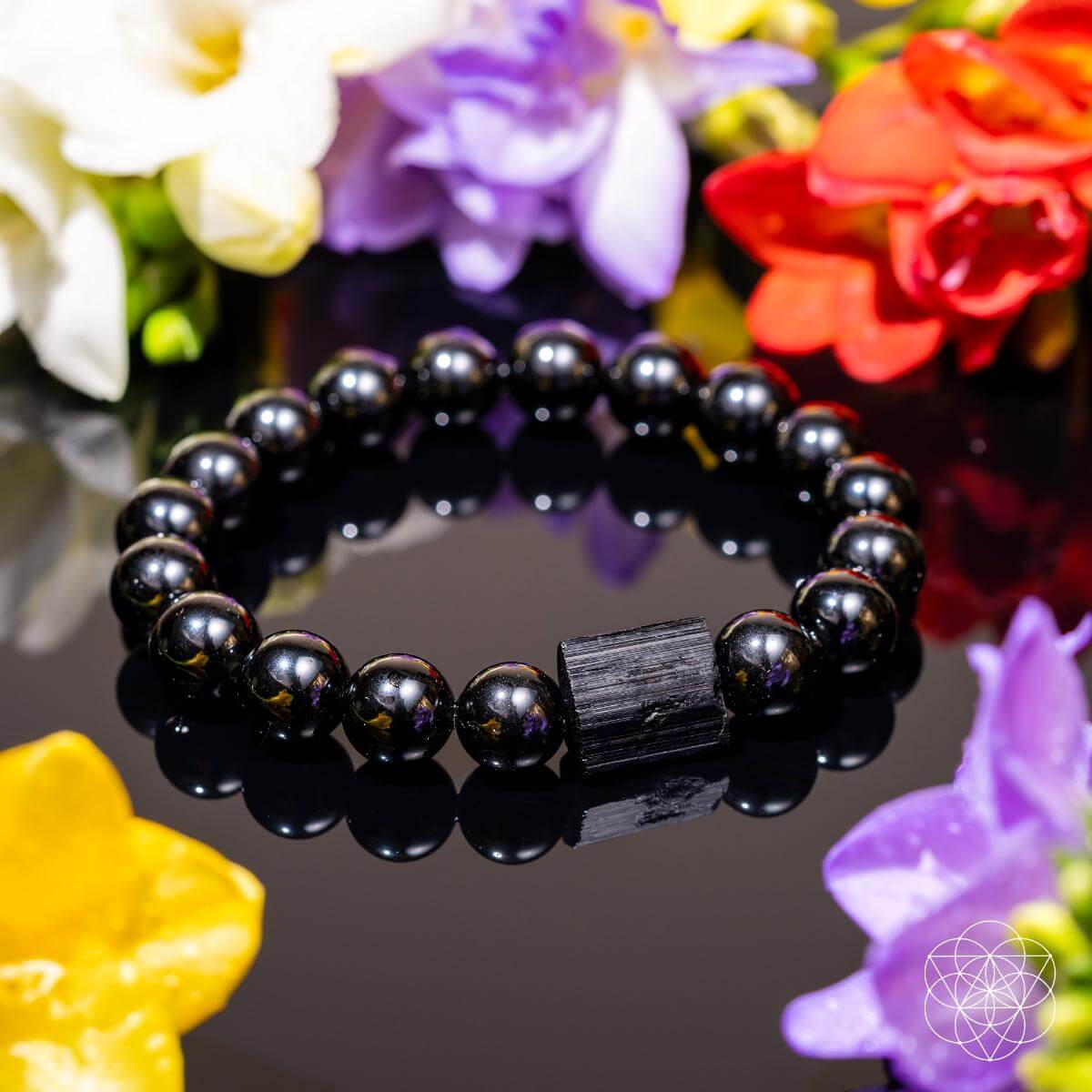 The Habit Breaker - Black Obsidian & Tourmaline Bracelet by Conscious Items features a rectangular central bead and is shown on a glossy black surface with vibrant flowers, with the brand’s geometric logo in the corner.