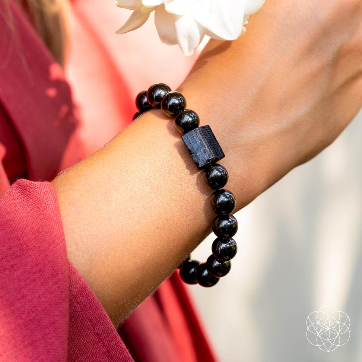 A close-up shows a wrist adorned with the Habit Breaker - Black Obsidian & Tourmaline Bracelet by Conscious Items, featuring a rectangular bead. The person wears a pinkish-red sleeve and holds a white flower against a soft background.