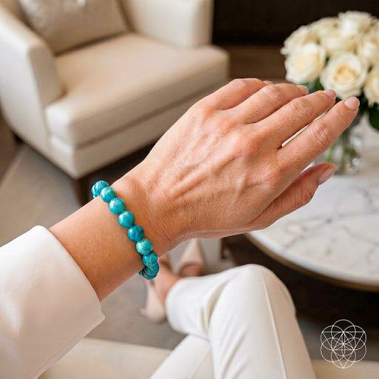 A person wearing the Empire of Her Own – Premium Blue Amazonite Bracelet by Conscious Items, inspired by Amazon Warriors, rests their hand on their knee. The background features a cream armchair, a white marble table, and white roses.