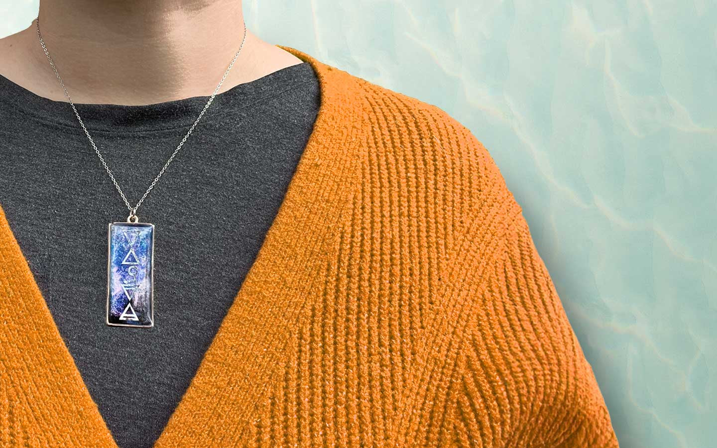 A person wearing a dark gray shirt and an orange knit cardigan displays a rectangular necklace with a blue and white geometric design, standing in front of a light, aqua-colored background.