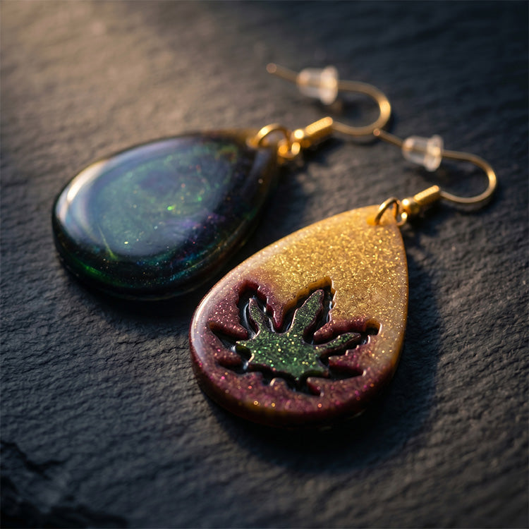 Teardrop resin earrings with gold, purple, and green glitter featuring an embedded leaf design on a dark stone surface.