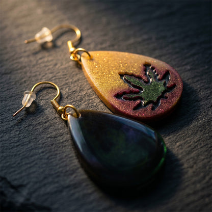 Close-up of asymmetrical resin earrings with gold, purple, and green glitter accents on a dark stone background.